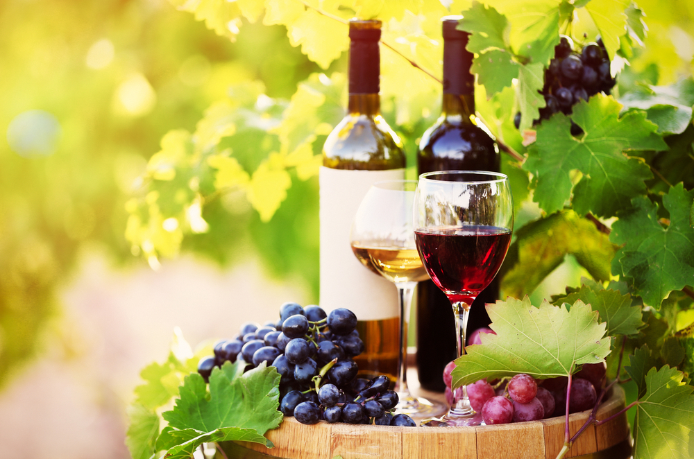 Two wine bottles and two wine glasses sitting on top of a barrel along with grapes.