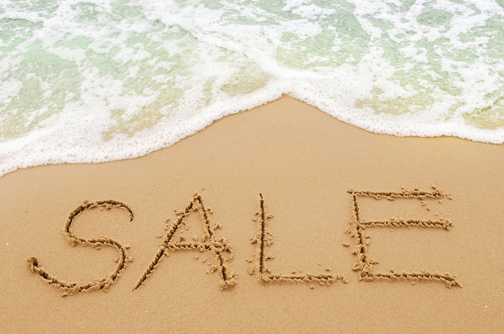 The word sale is written in the sand on a beach with ocean water running up to it.