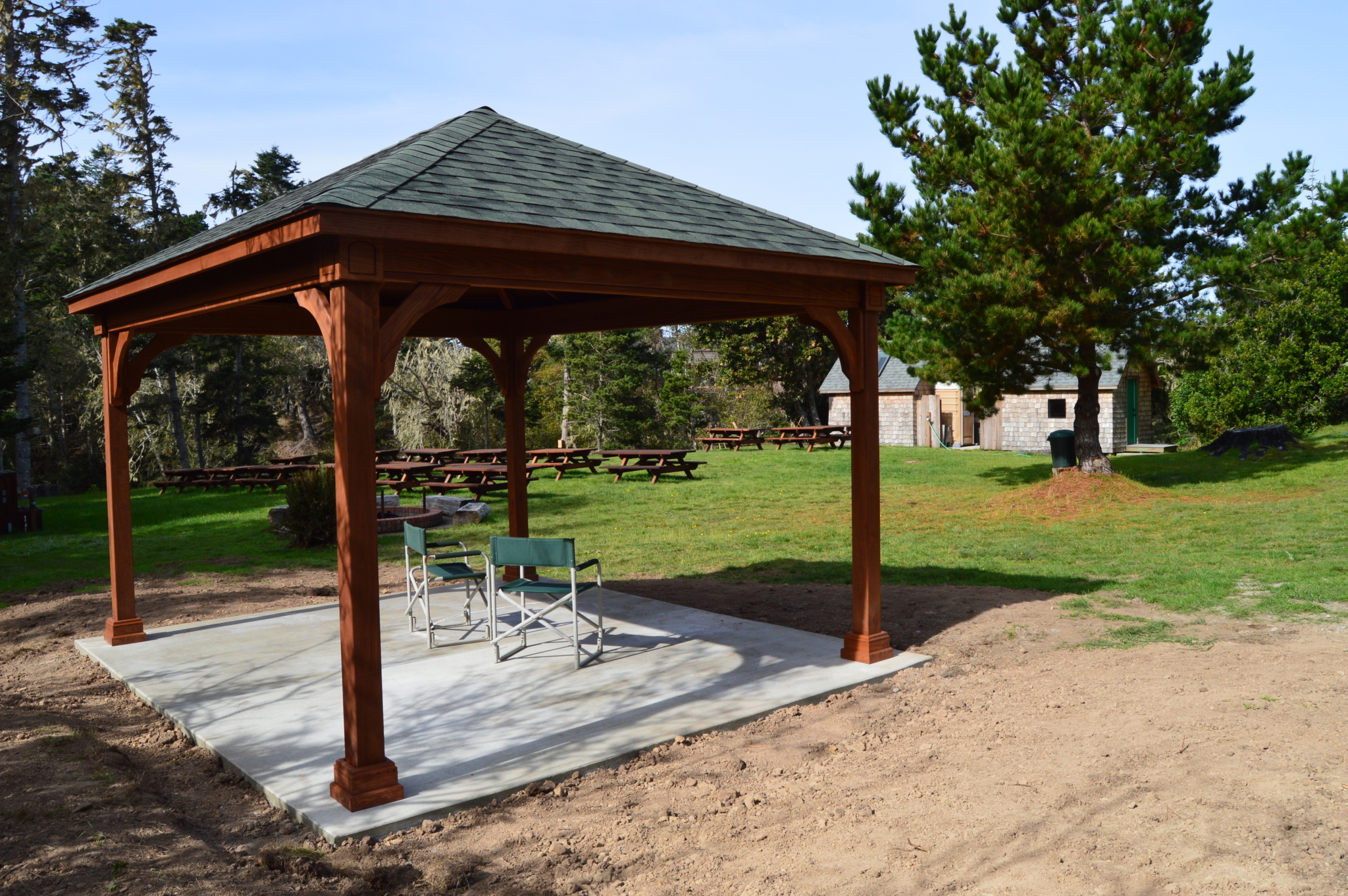 Small park shelter with two chairs under it and picnic tables in background